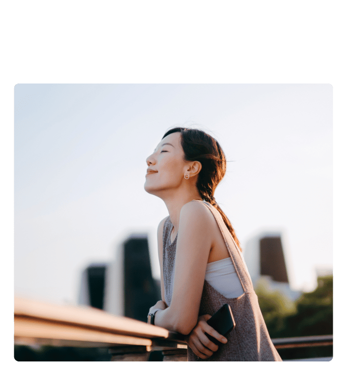 Person leans on a railing with eyes closed in warm sunlight against a city skyline