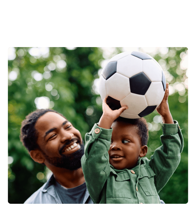 Smiling adult watches a child balance a soccer ball overhead among green trees