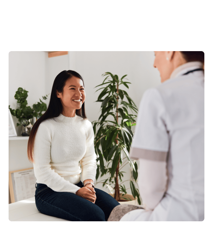 Smiling person in a white sweater sits on an exam table talking with a provider near potted plants