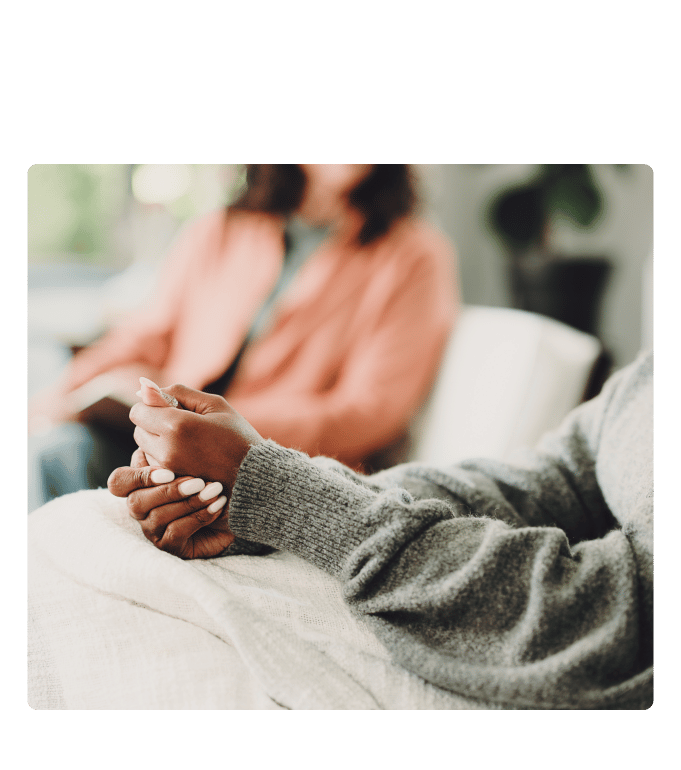 Person in a gray sweater clasps hands while talking with another person seated across the room