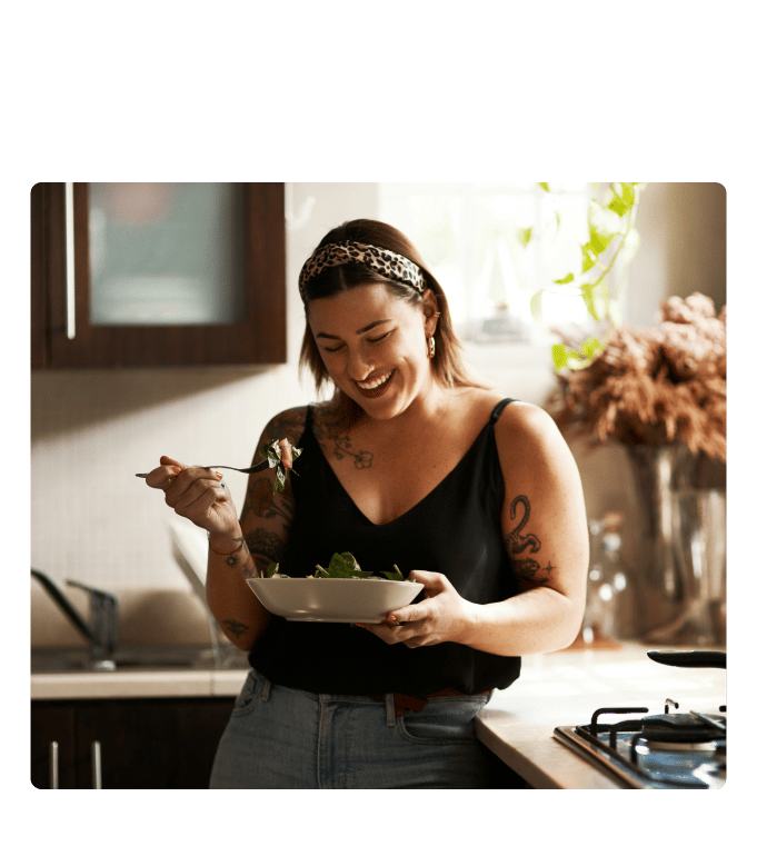 Smiling person in a leopard-print headband eats a bowl of salad in a kitchen