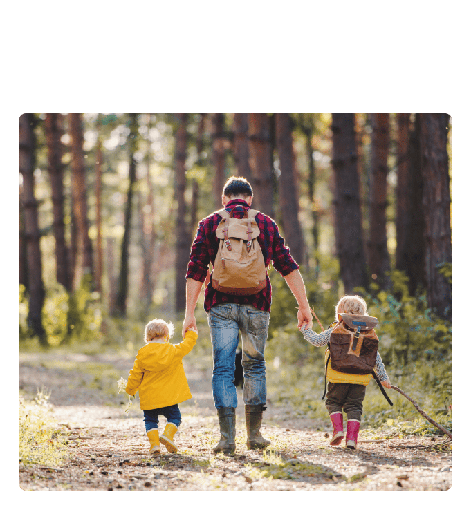 Adult and two children in rain boots walk hand in hand along a sunlit forest path