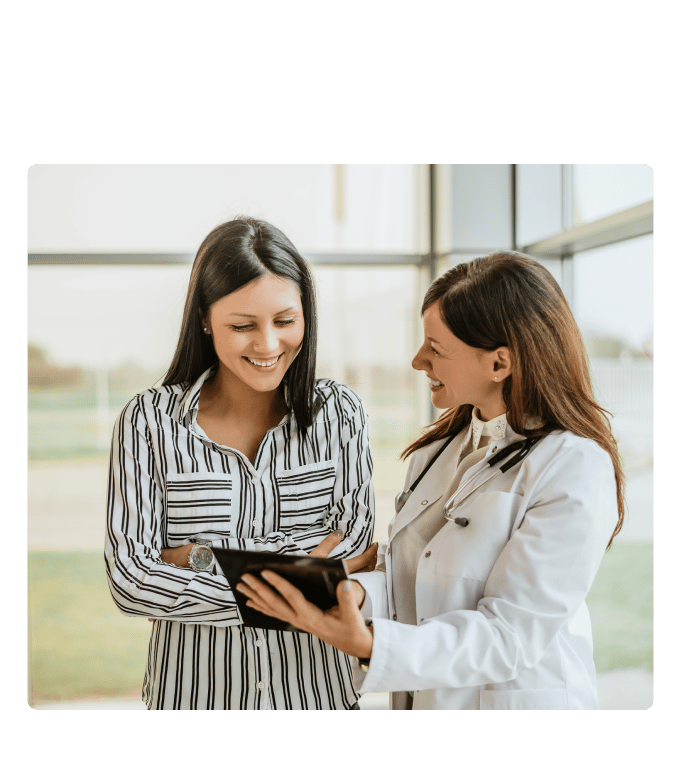 Provider in a white coat shows a tablet to a smiling person standing by tall windows