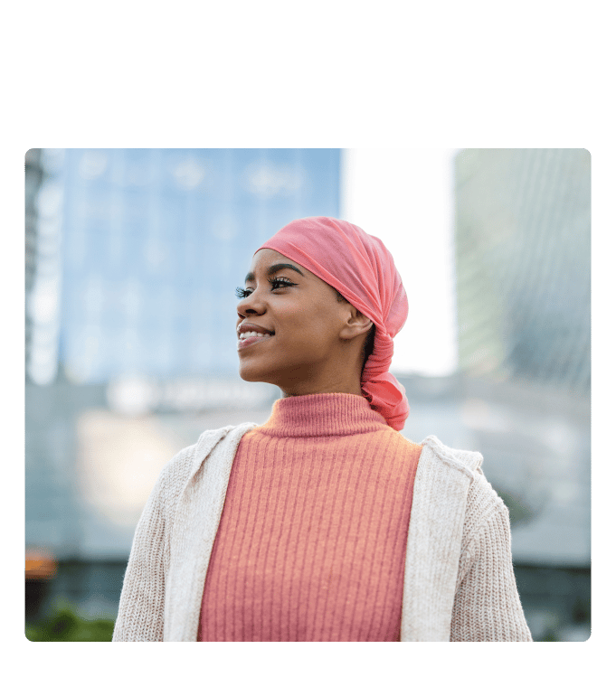 Smiling person in a pink head scarf and sweater standing outdoors among tall buildings