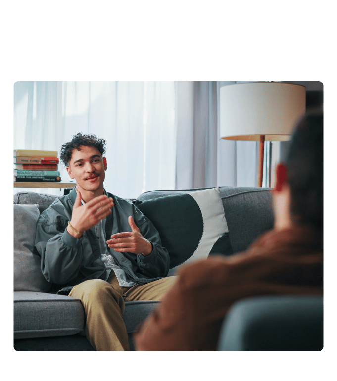 Person on a couch gestures while talking with another person in an armchair near a lamp