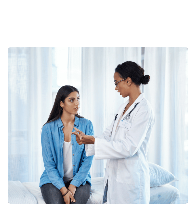 Healthcare provider in a white coat talks with a person seated on an exam bed by sheer curtains