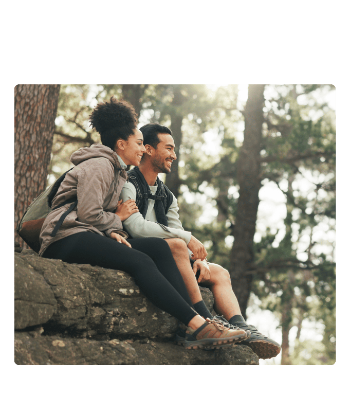 Two smiling people sit close together on a rocky ledge in a sunlit pine forest