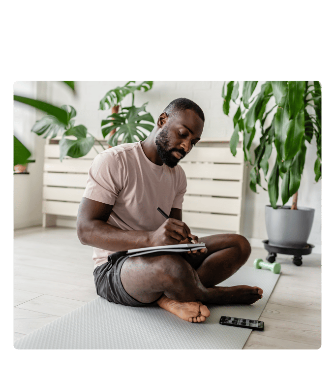 Person sitting cross-legged on an exercise mat writing in a notebook near houseplants and a dumbbell