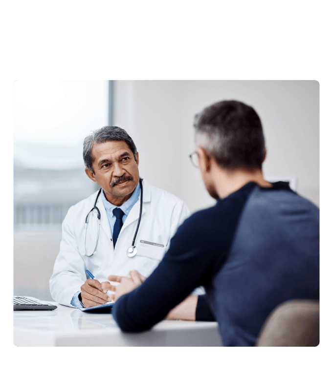 Doctor in a white coat with a stethoscope takes notes while talking with a person across a desk