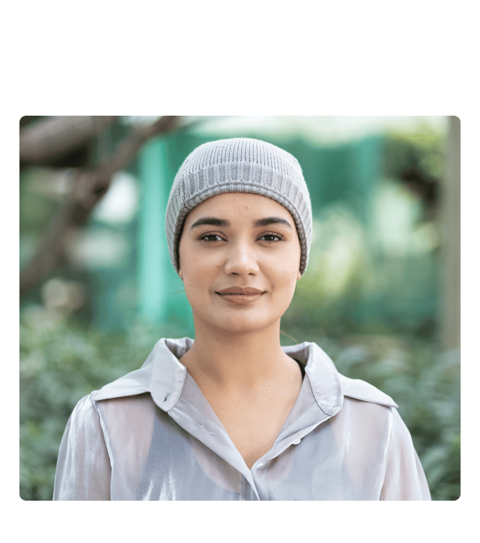 Smiling person in a gray knit beanie and shimmery blouse standing outdoors among greenery