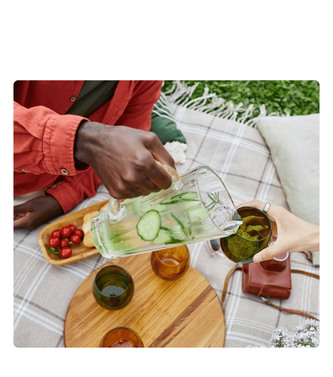 Person pours cucumber water into glasses on a picnic blanket with tomatoes and flowers nearby